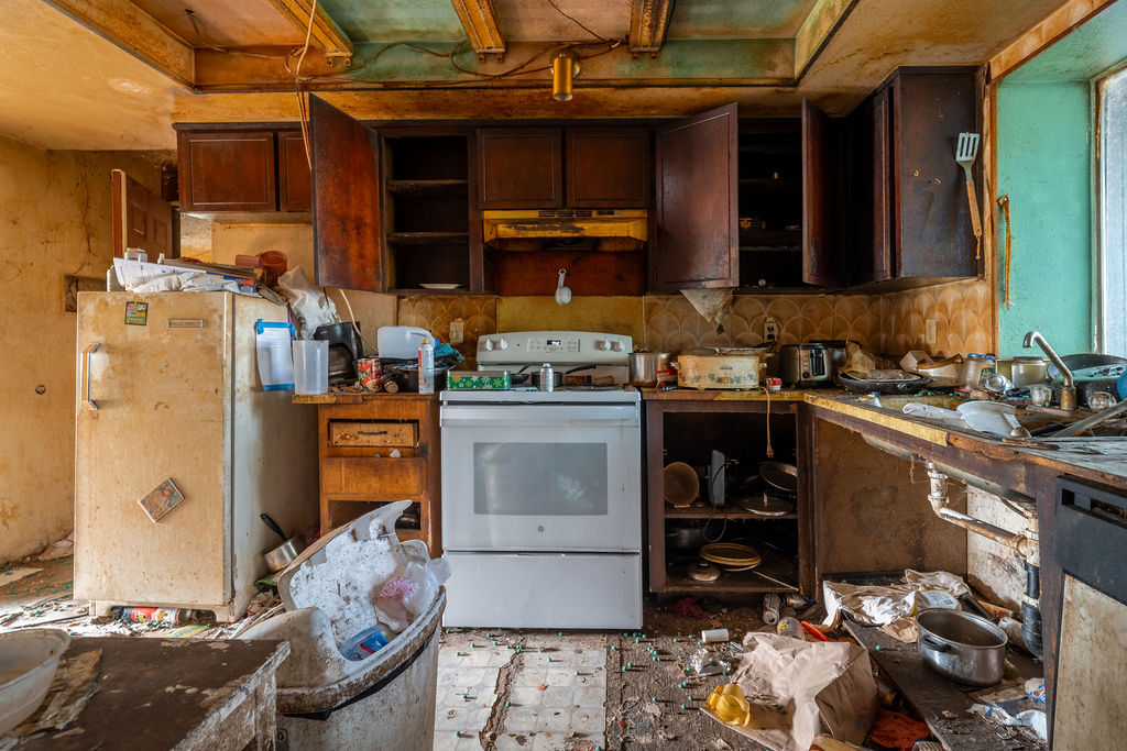Severely neglected kitchen with damaged cabinets, cluttered counters, and worn flooring before renovation